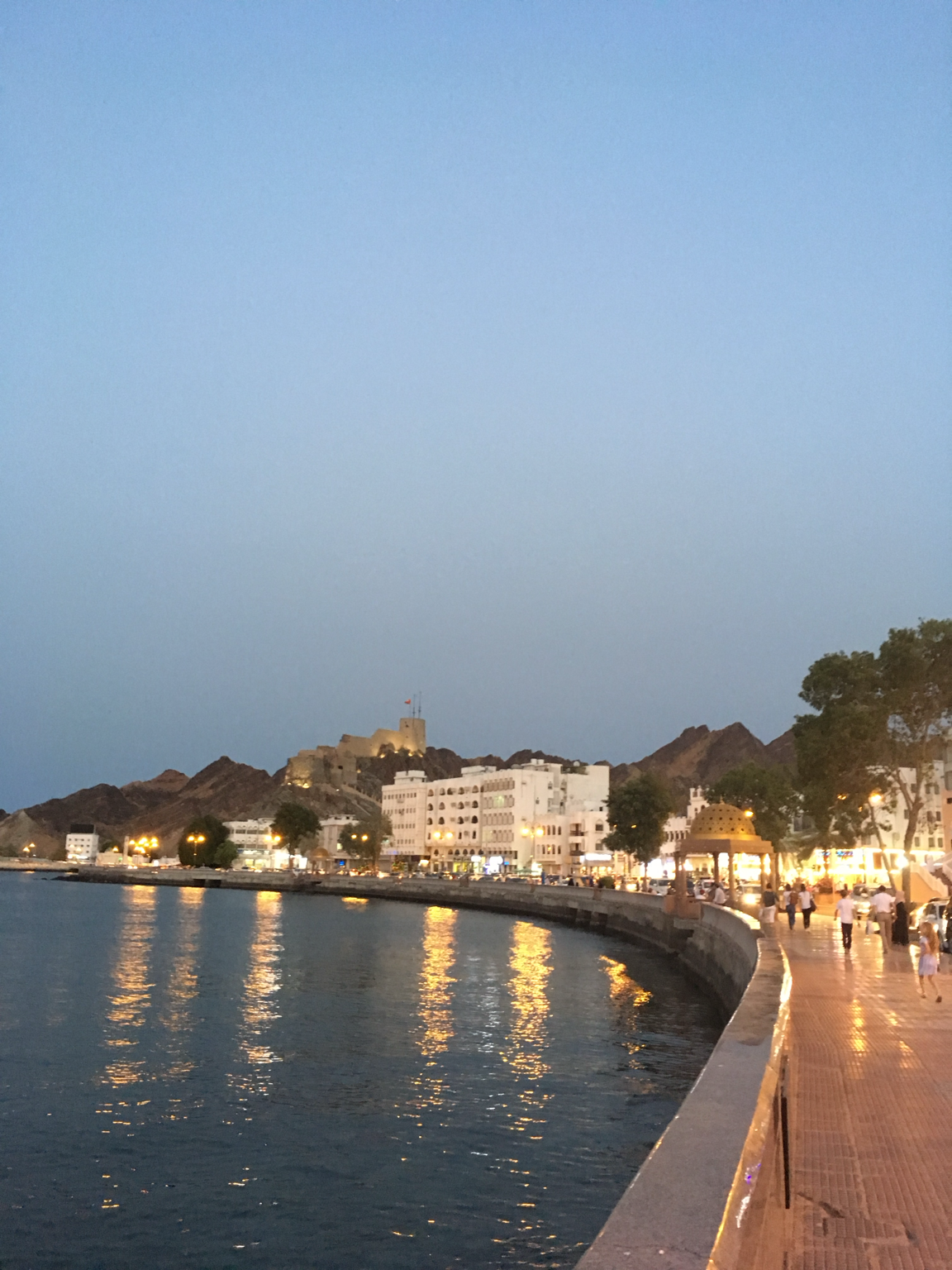 Illuminated Muttrah Corniche in Muscat, with waterfront buildings, mountains and reflections at dusk
